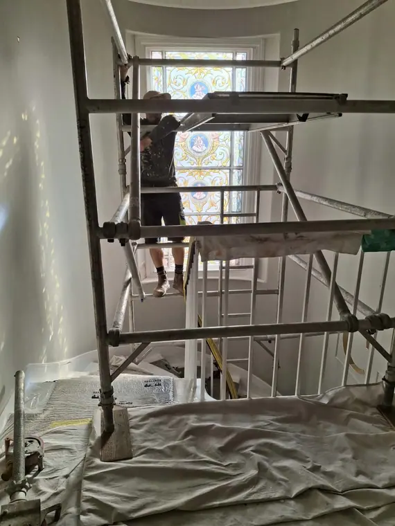 Working at full height in the stairwell beside the stained-glass window, with every surface sheeted.